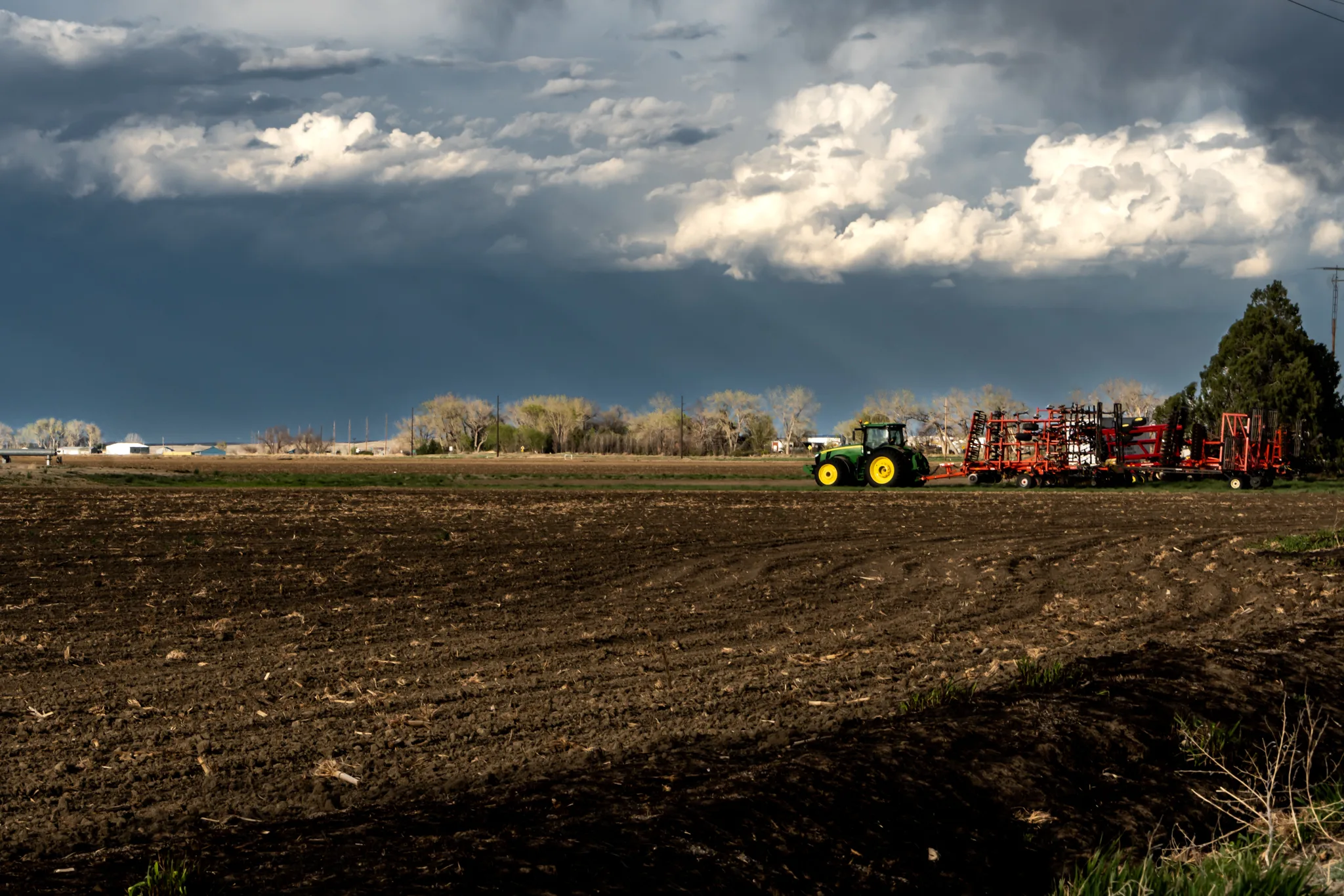 Westward view from eastern Morgan County Colorado of a tractor connected to implements, which are illuminated by a lowering sun against the backdrop of dark storm clouds that had just passed through.
