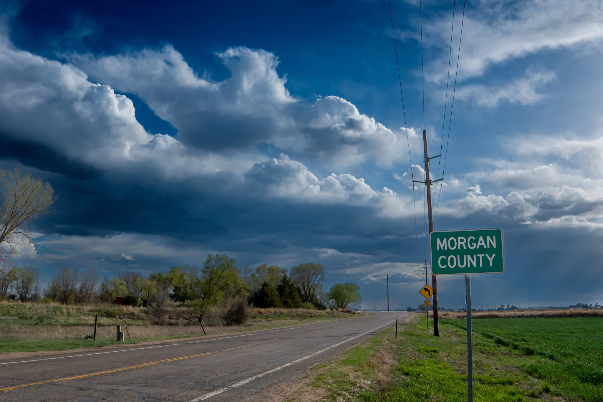 Green sign with white letters reading Morgan County, placed at the entrance to Morgan County Colorado as it sits along US Highway 6 looking eastbound, with large storm clouds growing in the background.